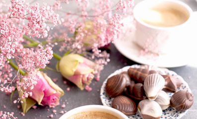 Fototapeta na wymiar A cup of fragrant coffee in the morning and a bouquet of pink flowers. Chocolate sweets in the form of cockleshells. Shells on the cups. Romance and light colors. Good morning. Card