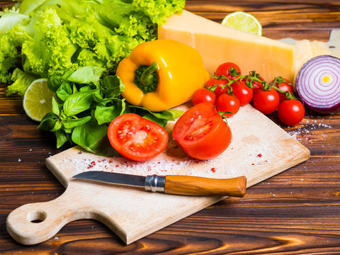 Appetizing still life. Fresh bulgarian yellow pepper, cherry tomatoes, cheese, lettuce leaves, red onion, green basil on wooden board. The knife for cutting vegetables. Top view