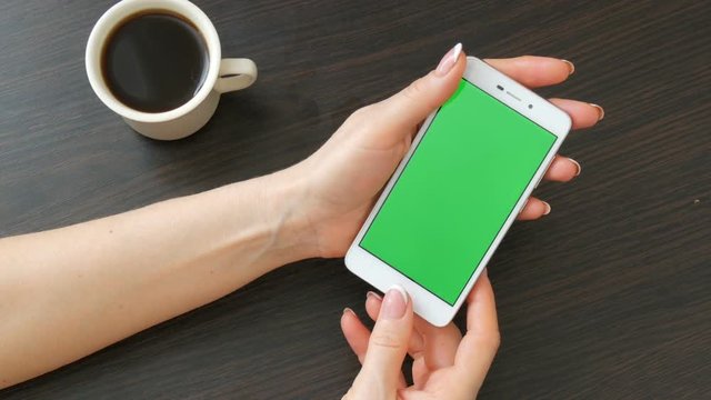 Female hands with beautiful French manicure take a white smartphone with Green Screen near white cup of coffee. Using Smartphone,Holding Smartphone with Green Screen on a stylish black wooden table