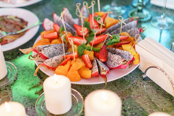 Fototapeta na wymiar Plate with sliced fruits and berries on a servered table. Wedding party in restaurant. Burning candle. White candles on glass candlesticks stand on tables at luxury wedding reception
