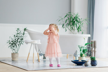 A little sweet girl stands in a large cozy light house near a white bathroom and closes her eyes with her hands with happiness. Playing hide and seek with parents