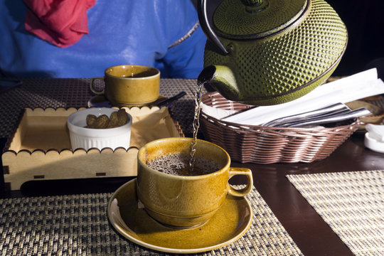 green tea from a old shabby cast iron kettle is poured into a cup closeup on a blurred background of the second full cup on the opposite side of the table