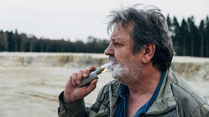 Old man with casual outfit is smoking an e-cigarette in forest area