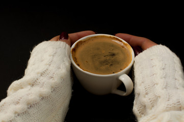 women's hands in a warm white sweater with a manicure hold a small white cup with coffee and a foam, Cozy mood
