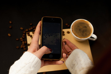 a woman's hand in a sweater photographing a cup of coffee on the phone and scattering coffee beans around the sheet empty space for the text of the text
