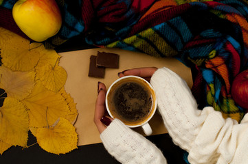 Woman's hand in a sweater holds a cup of coffee on the background of an envelope with fried leaves and colored apples, an autumnal mood