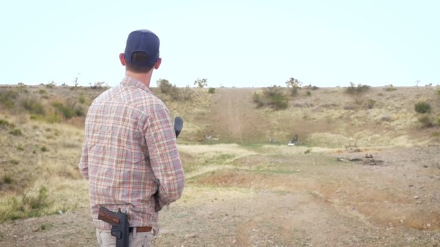 Wide shot of a man shooting a pump action shotgun at a desert gun range in slow motion. The young caucasian male wears a pistol on his belt as he fires his 12 gauge weapon at a clay pigeon.