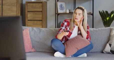 Young bored pretty blonde woman sitting on the gray couch with a remote control and changing channels in the living room. Inside - Powered by Adobe