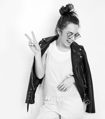 Portrait of young beautiful brunette woman girl model with nude makeup wearing summer hipster biker leather jacket clothes in sunglasses posing near wall. Showing her tongue and peace sign