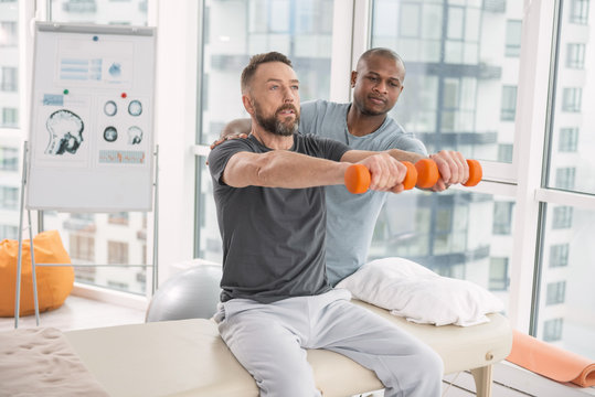 Rehabilitation exercise. Nice pleasant handsome man sitting on the medical couch and holding dumbbells in front of him