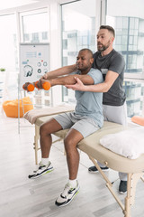 Medical therapy. Skillful handsome doctor standing behind his patient while helping him to lift dumbbells
