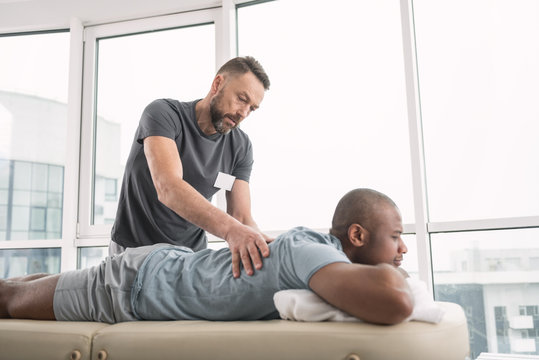 Experienced doctor. Serious skilled man looking at his patients back while doing a professional massage