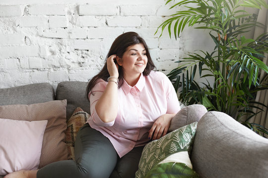 Cute young chubby brunette woman dressed casually having rest on large gray sofa, smiling shyly, adjusting her dark hair. Portrait of plus size body positive female enjoying leisure time indoors