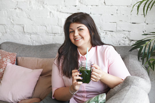 People, health, nutrition, diet and detoxication concept. Picture of happy cheerful young obese female wearing pink shirt having healthy green smoothie or shake for breakfast in the morning
