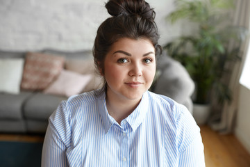 Beauty and body positivity concept. Picture of happy attractive young brunette plus size woman with hair bun and beautiful features posing indoors against gray couch and plant pot background - Powered by Adobe