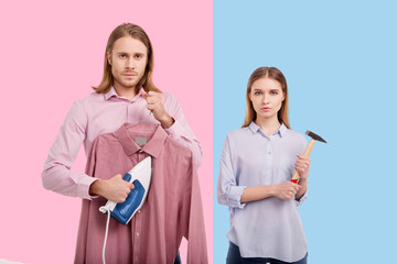 Ruining stereotypes. Handsome young man ironing a shirt and his girlfriend posing with a hammer...