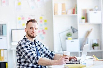 Portrait of young handsome bearded man in checked shirt looking at camera while sitting at laptop with coffee in modern office