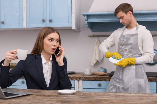 Terrible news. Shocked determined woman discussing work on the phone and her husband washing the dishes