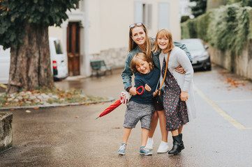 Young happy mother with two kids, little boy and girl, playing outside on a fresh rainy day. Stylish family having fun outdoors