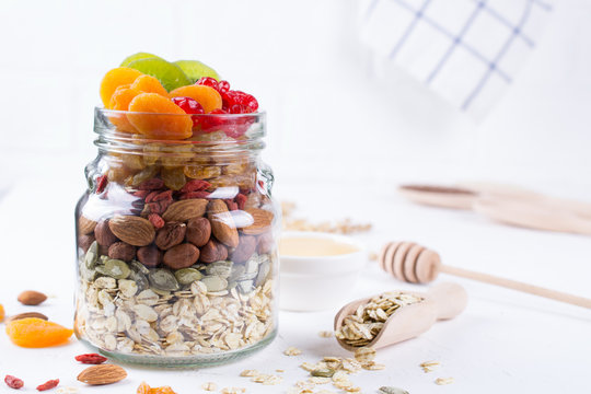 Glass jar with ingredients for cooking granola on white background. Oat flakes, honey, nuts, dried fruit and seeds. Healthy snak
