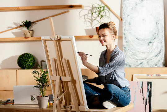 smiling female artist sitting on table and drawing on canvas in studio