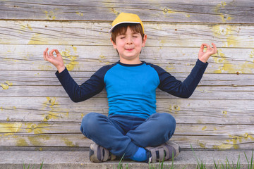 Portrait of a cute cheerful boy with yellow cap sitting outdoors in yoga pose having fun