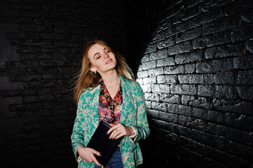 Stylish blonde girl in jacket and jeans with notebook diary at hands against brick black wall at studio.