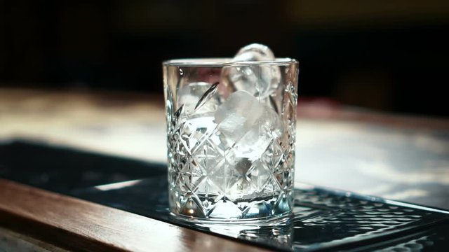 Barman pours pieces of ice into glass on table in night bar indoors. Young employee throws frozen chunks into transparent glass on desk in trendy place. Talented bartender prepares to make delicious