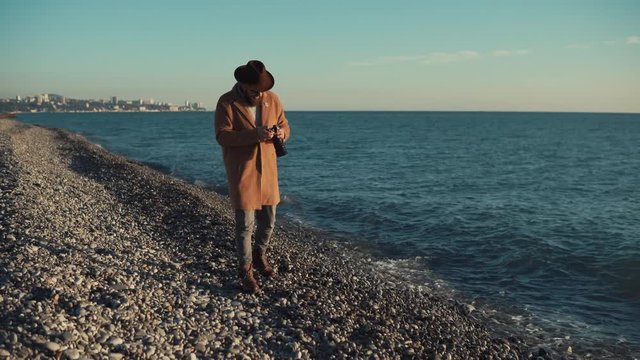 Stylish man is walking on the beach with a camera wearing warm clothes. He is taking pictures of sea and beach.