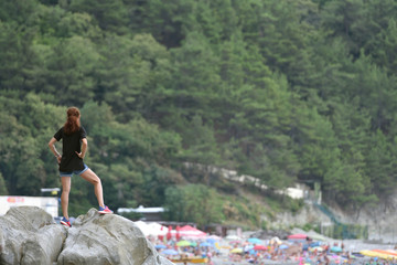 Naklejka premium Beautiful girl or woman is standing on a big stone and is looking at the beach with people