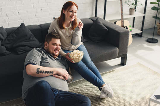 Smiling Overweight Boyfriend And Girlfriend Eating Popcorn And Watching Tv At Home