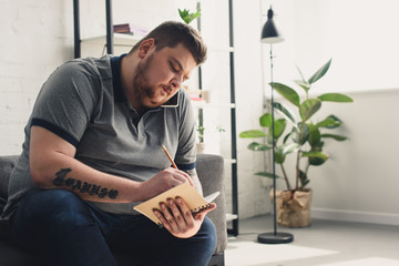 Handsome overweight man writing something to notebook on sofa at home - obrazy, fototapety, plakaty