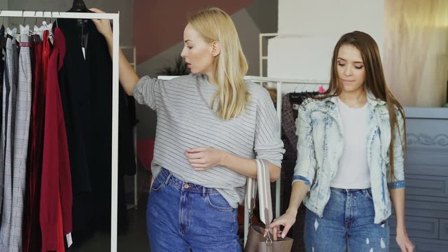 Young woman is stealing wallet from female customer while her victim is choosing clothes and not paying attention to her bag. Thief is looking around and going away.