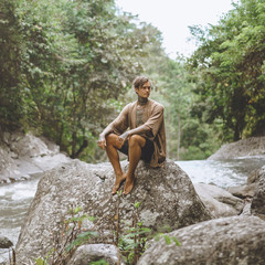 tattooed man resting on rock with green plants and river on backdrop, Bali, Indonesia