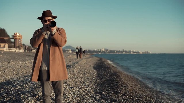 Happy adult man with thick beard holding a professional camera on the beach. He is taking picture of blue sea.