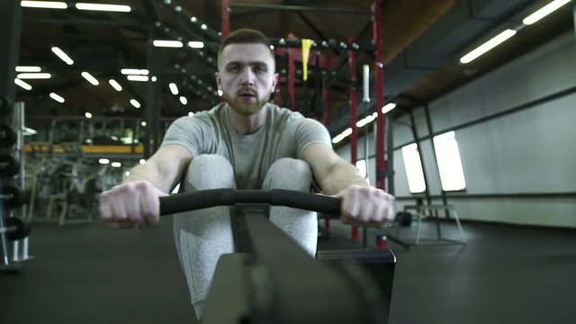 In gym on a rowing machine man performs an exercise in workout. A strong male athlete with an effort performs loads in hall for crossfire.