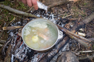 Old vintage pot in black soot is standing on the open fire in the wood. Natural summer holidays photo. Fish broth cooked in the forest. Vegetarian carrot and potatoes soup. Man holding salt cellar.