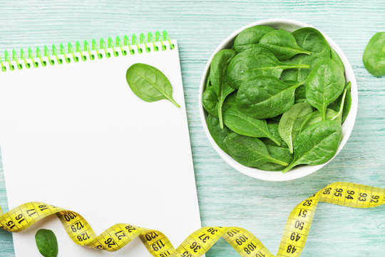 Notebook, green spinach leaves and tape measure on wooden table top view. Diet and healthy food concept.