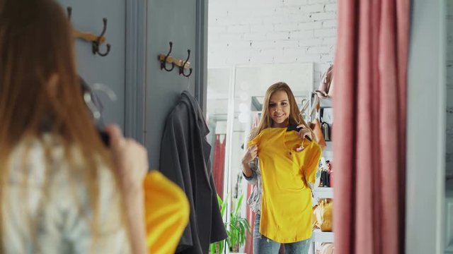 Mirror shot of young lady choosing clothes in fitting room. Girl is trying top and jumper, checking their size and length while standing opposite large mirror.