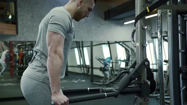 Portrait of young man, who is doing chest version of dips exercise in modern gym. Handsome sportsman in grey t-shirt is holding hangles of equipment and pushing his healthy muscular body up and down