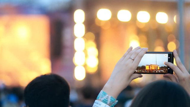 Young woman is photographing of music concert, standing outdoors in summer. Female fan takes photo of holiday event, using smartphone, holding device in hands. Scene, bright lights and watching people