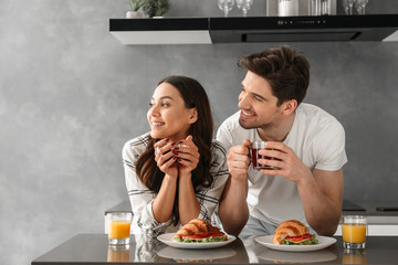 Young and beautiful couple looking aside and smiling, while having breakfast and drinking tea in flat