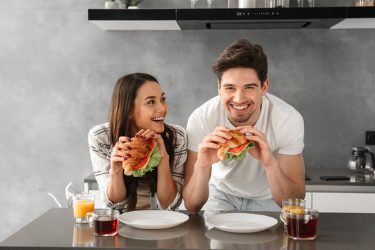 Young And Beautiful Couple Smiling, And Eating Sandwiches On Breakfast In Kitchen