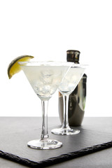 close up view of alcohol margarita cocktails with lime and shaker on grey surface on white