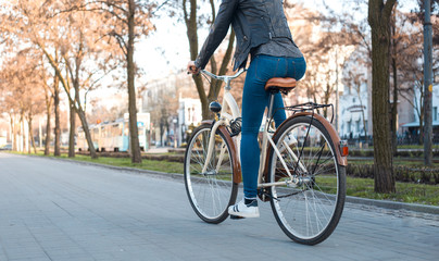 A girl in a leather jacket and jeans rides a bicycle along the avenue in the spring