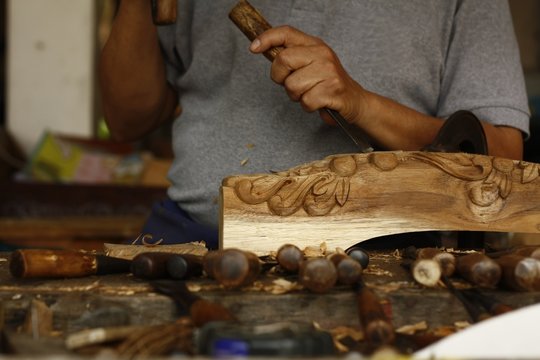 self taught skilled Thai man carving an intricate decoration on a hardwood table leg in his workshop using hand tools, Southeast Asia