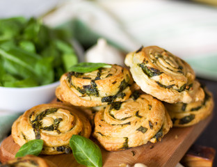 Buns of puff pastry with spinach and garlic close-up on a wooden board against a background of a bowl of spinach. Homemade pastries with greens.