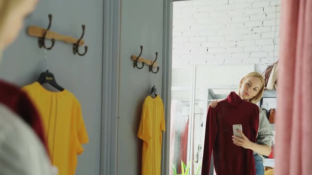 Female customer is making mirror selfie with new knitted jumper using smart phone. She is standing in fitting room in clothes store and posing with lovely smile.