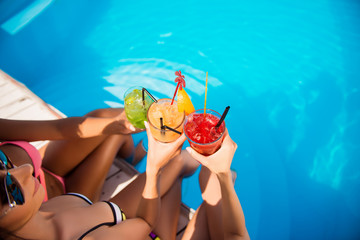 High angle cropped shot of three girls cheering with drinks near swim pool. Red, green and yellow...