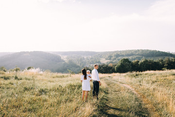 Naklejka na ściany i meble Walk beautiful young family in white clothes with a young son blond in mountainous areas with tall grass at sunset. Mother keeps son in her arms, hugging. family - this is happiness